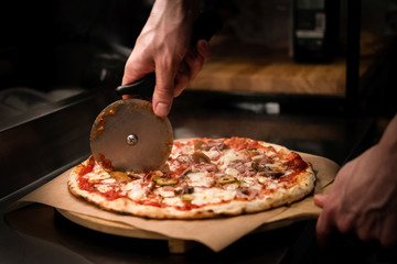 Chef cutting freshly baked delicious pizza with pizza cutter on professional kitchen. Close up of a...