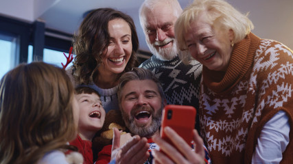 Happy extended family with grandparents making a fun self portrait together using mobile phone embracing together celebrating Christmas. Noel gathering.