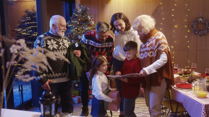 Beautiful big extended family gathering together on Christmas night. Cheerful little boy with parents, sister and grandparents reading magic Christmas story book together.