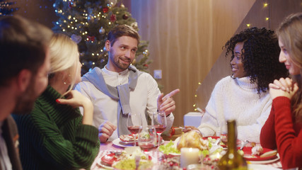 Christmas party. Happy attractive interracial guys and ladies talking at dining table eating food talking celebrating winter holidays.