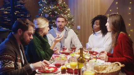 Christmas happy young people dining together at holiday table. Blonde pretty girl telling a fantastic surprising story to her friends. Concept of communication.