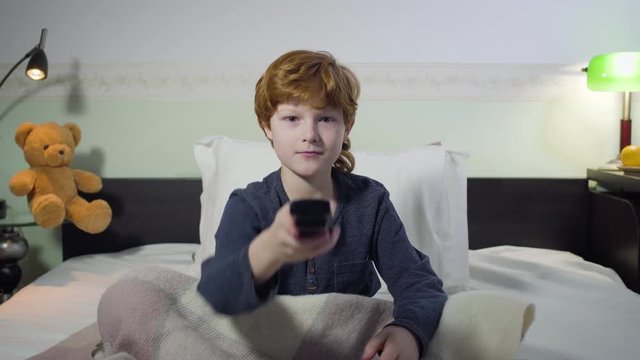 Front view portrait of cute Caucasian redhead boy sitting on bed and switching channels with remote control. Charming male child searching for interesting cartoons. Childhood, leisure, bedtime. - Powered by Adobe
