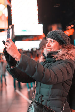 Beautiful curly brunette woman taking a selfie with her smartphone in Times Square, New york City. View over the shoulder, closeup