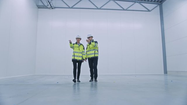 Two Engineers Talking, Walking Across Empty Factory Floor, inspecting, Planning Where All the Machinery Would go. Professionals Industrial Designers Strategizing about New Manufacturing Plant Design