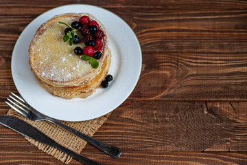 A stack of delicious pancakes with raspberries, blackberries and blueberries. On a wooden background. Sprinkled with powdered sugar and decorated with mint leaves.