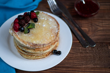 A stack of delicious pancakes with raspberries, blackberries and blueberries. On a wooden background. Sprinkled with powdered sugar and decorated with mint leaves.