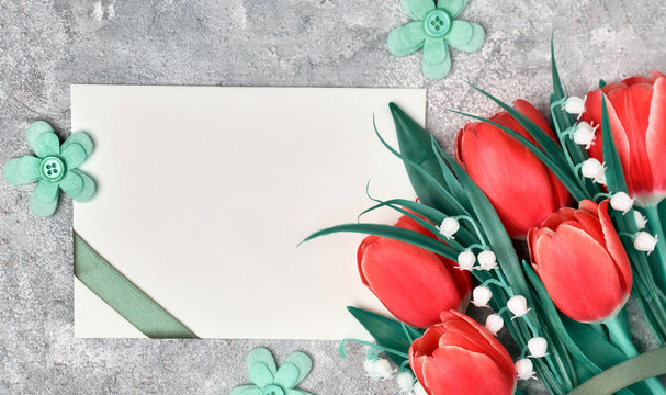 Spring greetings, flat lay with red tulips and blank paper card on grey textured background. Easter, Mother's day, Birthday or Anniversary, gender neutral greeting design.