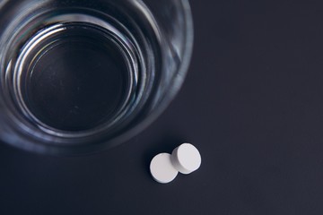 Two white pills on a dark background and a glass of clear water