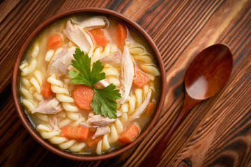 Top view close-up of a bowl of chicken noodle soup with a wooden spoon on a wooden rustic background