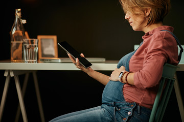 Side view of cute smiling caucasian pregnant woman sitting in home office and using tablet.