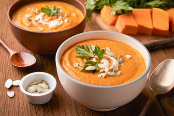 Two bowls of pumpkin soup sprinkled with peeled pumpkin seeds and a raw pumpkin with parsley on a cutting board
