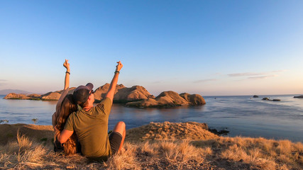A couple sitting on top of a small island, enjoying the morning sun over Komodo National Park, Flores, Indonesia. Golden hour over the islands and sea. They form a heart with theirs hands. Love