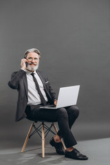 Fototapeta premium Business and fashionable bearded mature man in a gray suit taking by phone and using laptop on the grey background.