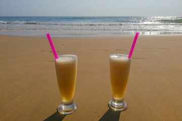 Two glasses with drinks and a pink straw stand on the sand on the beach, by the sea.