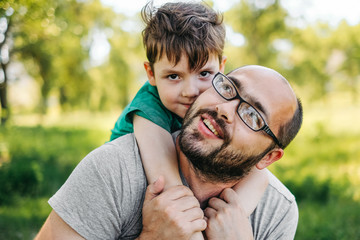 Fototapeta premium Portrait of handsome dad with a beard, looking at camera and smiling, hugging his cute little boy, trees in the background