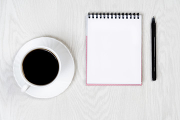 White cup with coffee on a white table with a notebook and pen