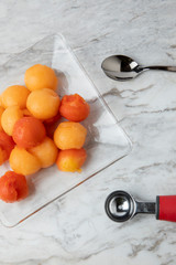 Bolitas de fruta en un plato de cristal, sobre una barra de mármol blanco con una cuchara y el cortador