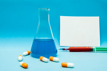 flask with a blue liquid, orange-white capsules, a syringe with blood on a blue background, in the background a white sheet of paper for copy space