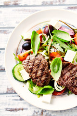 Grilled beef steaks with feta cheese, black and kalamata olives, capers, red onion and fresh basil. Bright wooden background. Top view. 