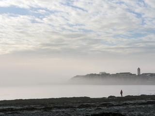 Galway bay and Grattan beach, Cloudy sky, Fog over houses, one person silhouette standing by the ocean, low tide.