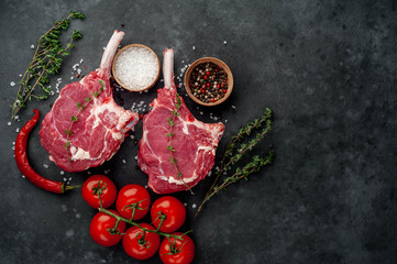 two raw steaks, beef tomahawk with spices, tomatoes and thyme on a stone background. with copy...