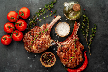  two grilled beef steak - tomahawk with spices, tomatoes, sunflower oil on a stone background.