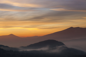 Valley filled with soft mist, distant silhouettes of impressive mountains and amazing colors of vivid morning sky
