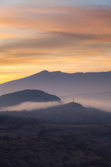 Soft, morning mist covering village houses, mountain layers and valley, distant pointy mountain peak and amazing vivid colors of sunrise sky