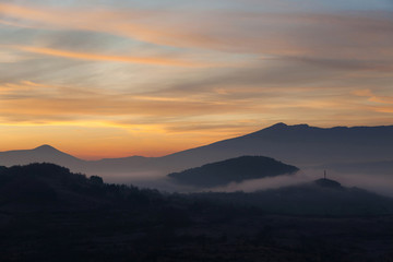 Soft, morning mist covering village houses, mountain layers and valley, distant pointy mountain peak and amazing vivid colors of sunrise sky