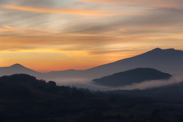 Valley filled with soft mist, distant silhouettes of impressive mountains and amazing colors of vivid morning sky