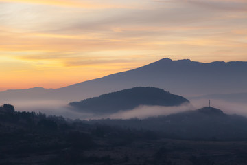 Soft, morning mist covering village houses, mountain layers and valley, distant pointy mountain peak and amazing vivid colors of sunrise sky
