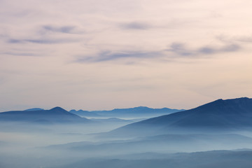 Pastel color palette of misty landscape with rocky mountain peak rising above fog covered valleys and mountain layers during colorful 