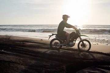 Fototapeta na wymiar Male masculine model with cross road motorbike at the ocean coast with black sand at the golden hour time