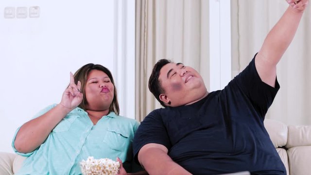 Happy obese couple taking selfie photo with a smartphone while sitting on the sofa in the living room at home. Shot in 4k resolution
