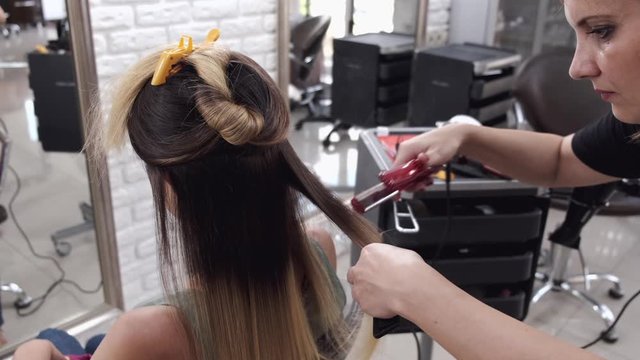 A client has long hair. She is sitting in a salon. The stylist is doing the hairstyle.