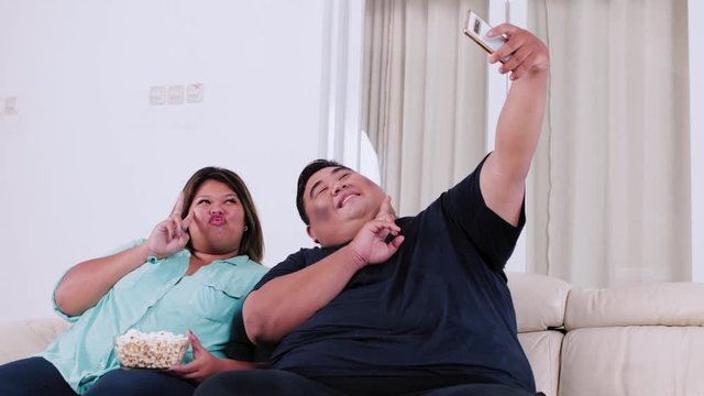 Happy obese couple sitting in the living room while taking selfie photo together with a mobile phone. Shot in 4k resolution