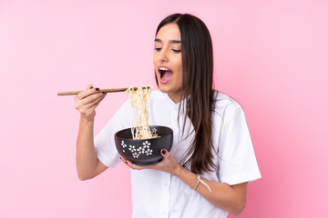 Young brunette woman over isolated pink background holding a bowl of noodles with chopsticks and eating it
