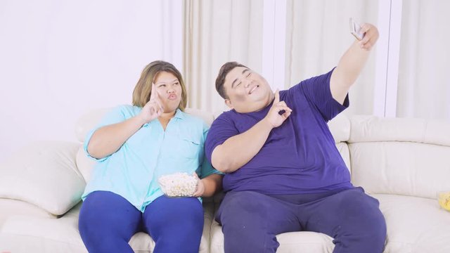 Obese couple using a mobile phone to take selfie photo while sitting on the sofa in the living room at home. Shot in 4k resolution