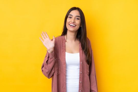 Young brunette woman over isolated yellow background saluting with hand with happy expression