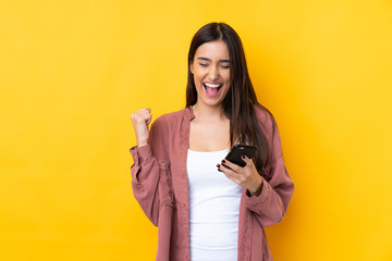 Young brunette woman over isolated yellow background with phone in victory position
