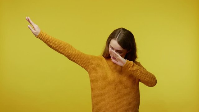 Happy positive girl with brown hair in pullover showing dab dance pose, famous internet meme of success, gesture of triumph, performing dabbing trends. indoor studio shot isolated on yellow background