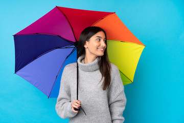 Young brunette woman holding an umbrella over isolated blue wall laughing