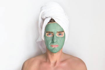 Young handsome man, metrosexual guy with cosmetic clay blue mask on his face and a towel on head...