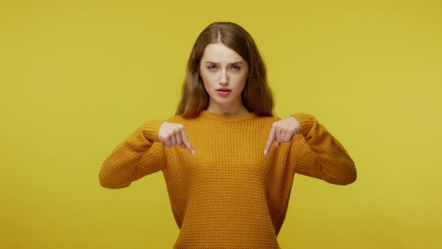 Strict bossy young woman in casual clothes pointing finger down and looking angrily at camera, demanding to be immediately here, control concept. indoor studio shot isolated on yellow background