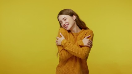 Beautiful lovely happy girl with brown hair in pullover embracing herself and smiling with dreamy satisfied expression, love yourself concept. indoor studio shot isolated on yellow background
