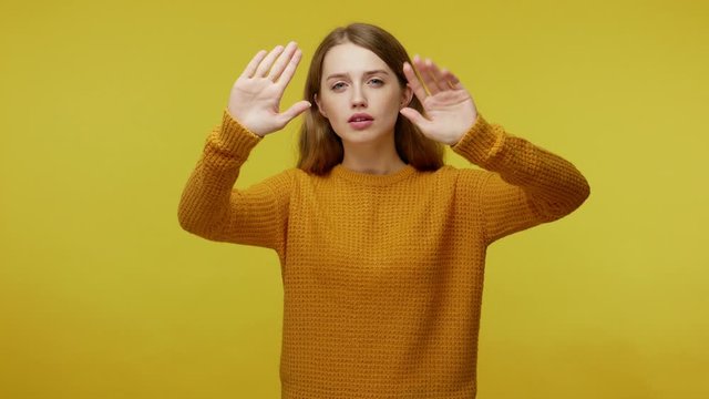 Attentive serious girl in pullover touching invisible wall or pushing buttons on screen with her palms, looking with interest, showing pantomime. indoor studio shot isolated on yellow background