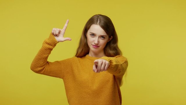 Displeased girl expressing disrespect, making L sign and pointing to camera saying you lost, showing looser or lame gesture, accusing for failure. indoor studio shot isolated on yellow background