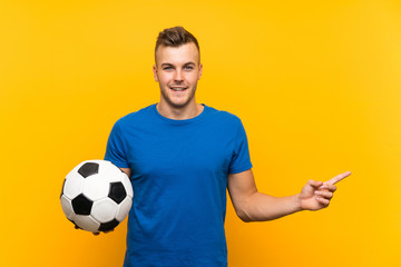 Young handsome blonde man holding a soccer ball over isolated yellow background surprised and...