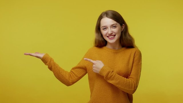 Smiling cute happy girl with kind face expression in pullover pointing copy space on palm against yellow background, showing advertising area for idea or image. indoor studio shot, workspace mock up
