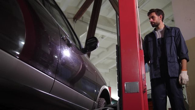 Auto repair specialist raising car with special lift while going to check chassis during vehicle maintenance. Handsome bearded repairman lifting car to see bottom for inspection in auto repair service